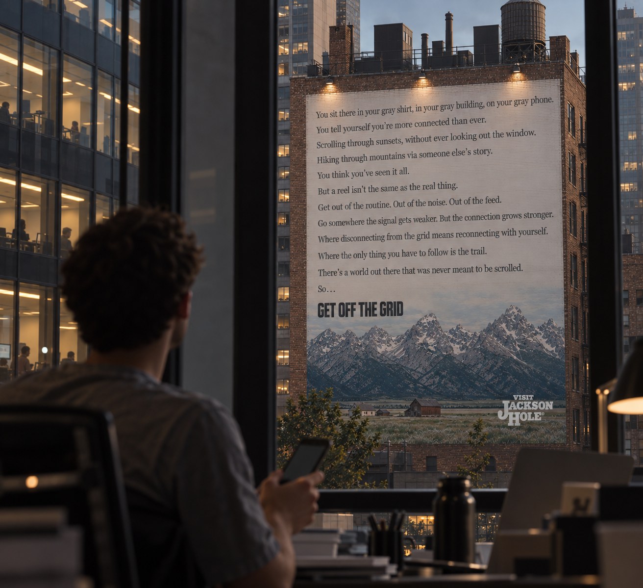 A building-side mural execution of the Visit Jackson Hole 'Get Off the Grid' campaign, seen through an office window as someone scrolls on their phone
